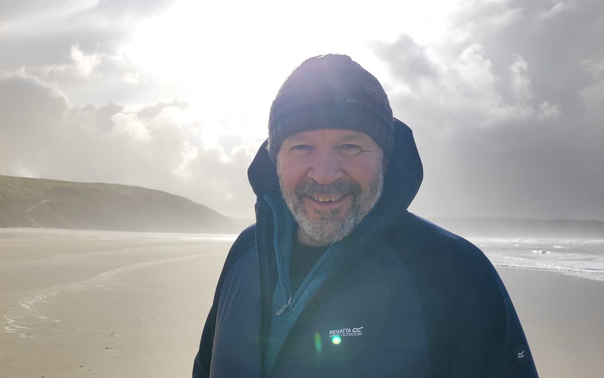 Ian smiling on a wide, empty beach, with the sun breaking through cloud behind him